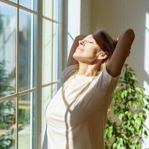 Mulher feliz espreguiçando o corpo e sorrindo de olhos fechados sob os raios de sol entrando pela janela numa manhã ensolarada em casa. Conceito de otimismo e felicidade