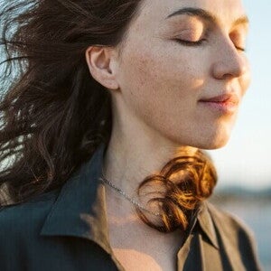 Mulher calma com cabelos esvoaçando ao vento na praia, sol no rosto.