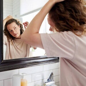Mulher em frente ao espelho passando os dedos pelo cabelo, verificando a queda de cabelo.