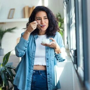 Mulher feliz comendo iogurte alimento com calcio enquanto está em pé na sala de estar em casa.