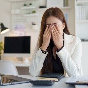 Mulher jovem em um escritório com as mãos no rosto e olhos fechados, expressando exaustão e estresse em frente ao notebook.