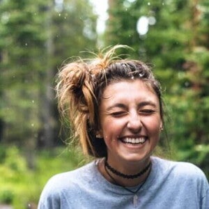 Mulher jovem sorri com olhos fechados, flocos de neve no cabelo, fundo verde desfocado.