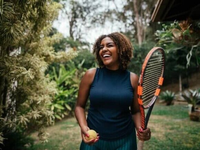 Mulher sorrindo com raquete e bola de tênis, representando energia e vitalidade.