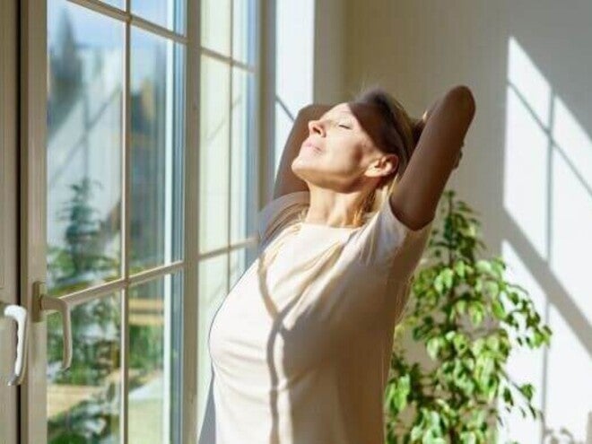 Mulher feliz espreguiçando o corpo e sorrindo de olhos fechados sob os raios de sol entrando pela janela numa manhã ensolarada em casa. Conceito de otimismo e felicidade