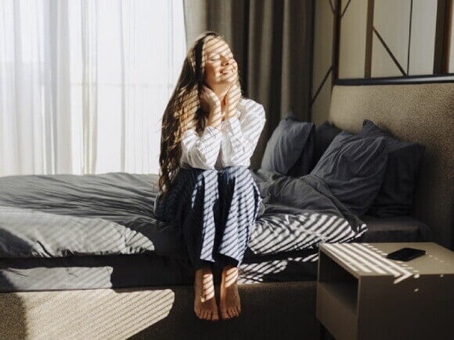Mulher sentada na cama, sorrindo e se espreguiçando depois de acordar.
