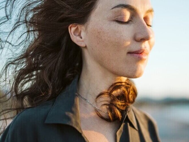 Mulher calma com cabelos esvoaçando ao vento na praia, sol no rosto.