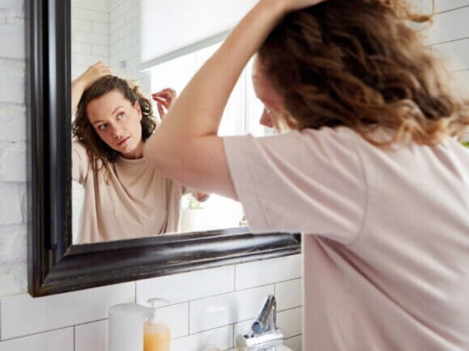 Mulher em frente ao espelho passando os dedos pelo cabelo, verificando a queda de cabelo.