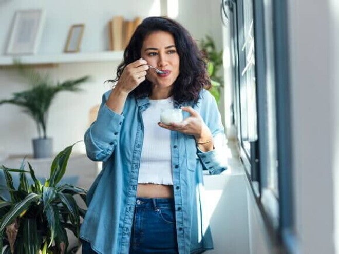 Mulher feliz comendo iogurte alimento com calcio enquanto está em pé na sala de estar em casa.