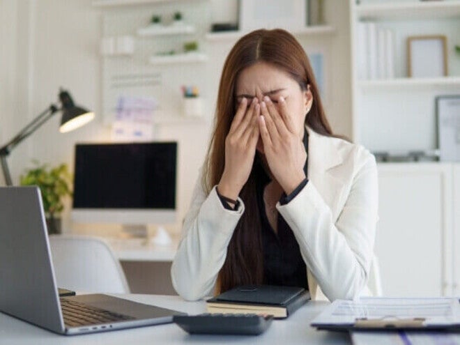 Mulher jovem em um escritório com as mãos no rosto e olhos fechados, expressando exaustão e estresse em frente ao notebook.