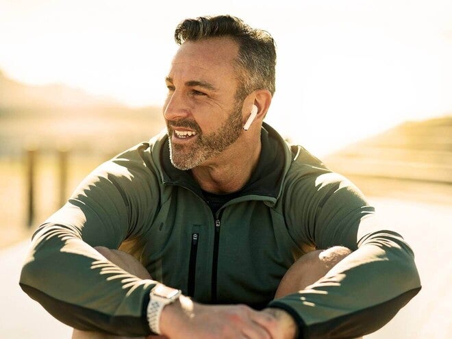 Homem adulto sorrindo, sentado ao ar livre e ouvindo música sob o sol.