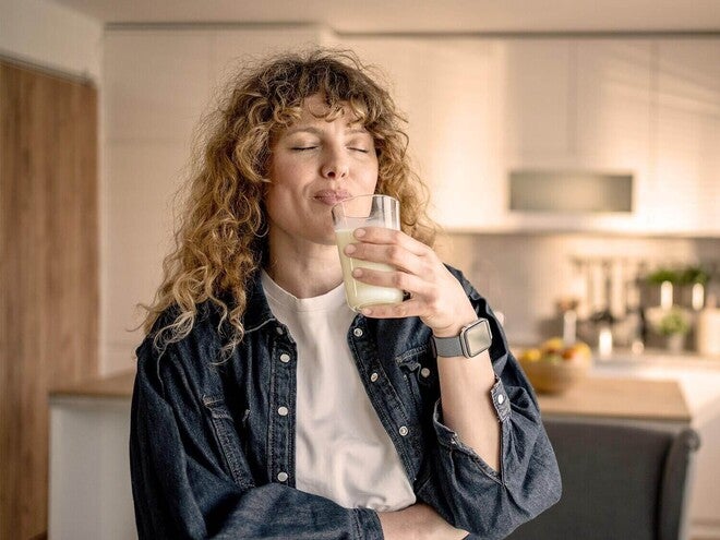 Mulher adulta em pé na cozinha, sorrindo e tomando um smoothie com Nestlé Vital Ritual Matinal