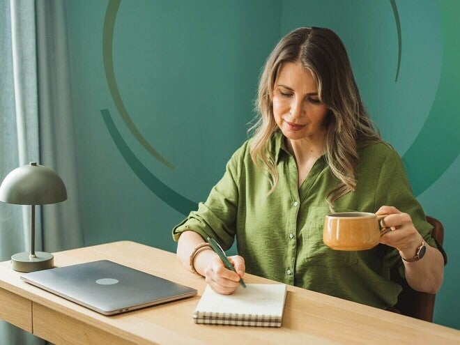 Mulher adulta sentada à escrivaninha, escrevendo em um caderno enquanto segura uma xícara de café