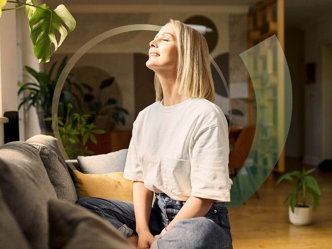 Mulher sentada no sofá de casa, sorrindo de olhos fechados enquanto sente a luz do sol entrar pela janela.