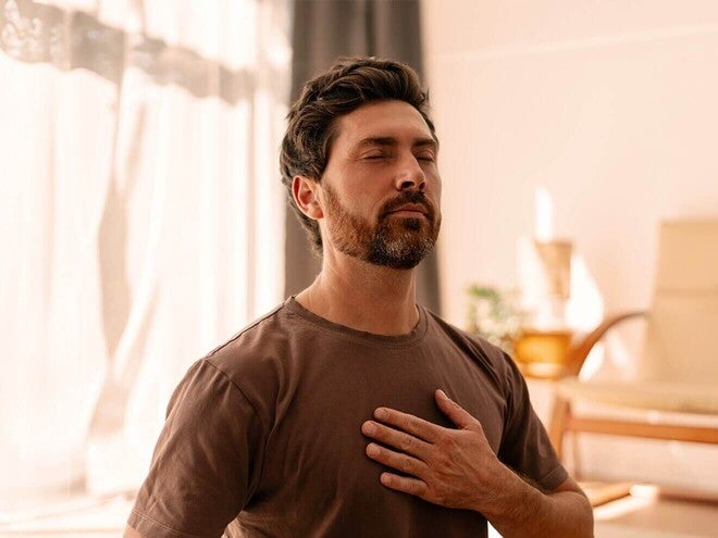 Homem adulto na sala de casa, de olhos fechados e com uma mão no peito, respirando com calma.