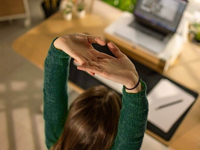 Mulher em frente ao computador, no escritório, alongando os braços para cima.