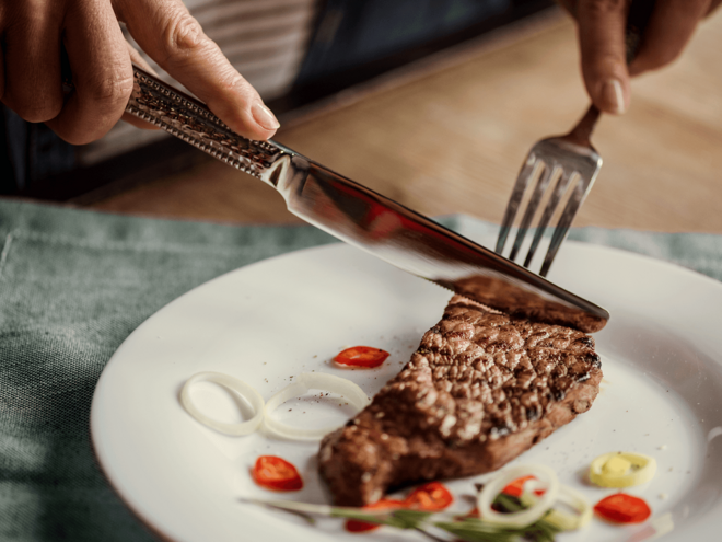 Pessoa cortando carne com faca e garfo. Comida com proteína.