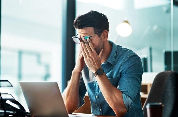 Homem no escritório, sentado em frente ao computador, estressado, cobre o rosto com as mãos