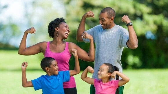 Uma família com dois adultos e duas crianças sorrindo e exibindo os músculos dos braços em um parque.