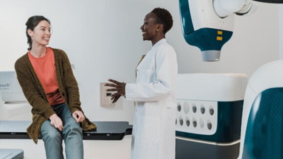 Médica sorridente de jaleco branco dialoga com paciente em maca em ambiente médico moderno.